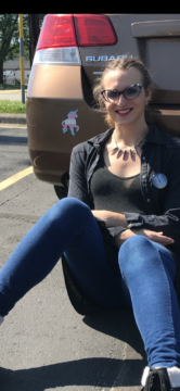 Brittyn sitting next to her car. She is wearing a "Trans Rights are Human Rights" button and there is a trans pride unicorn bumper sticker in the photo.
