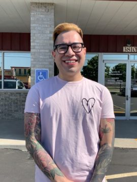 Tim is smiling in the Access to Independence parking lot, wearing a pink t-shirt with a heart on it. He has sleave tattoos and glasses.