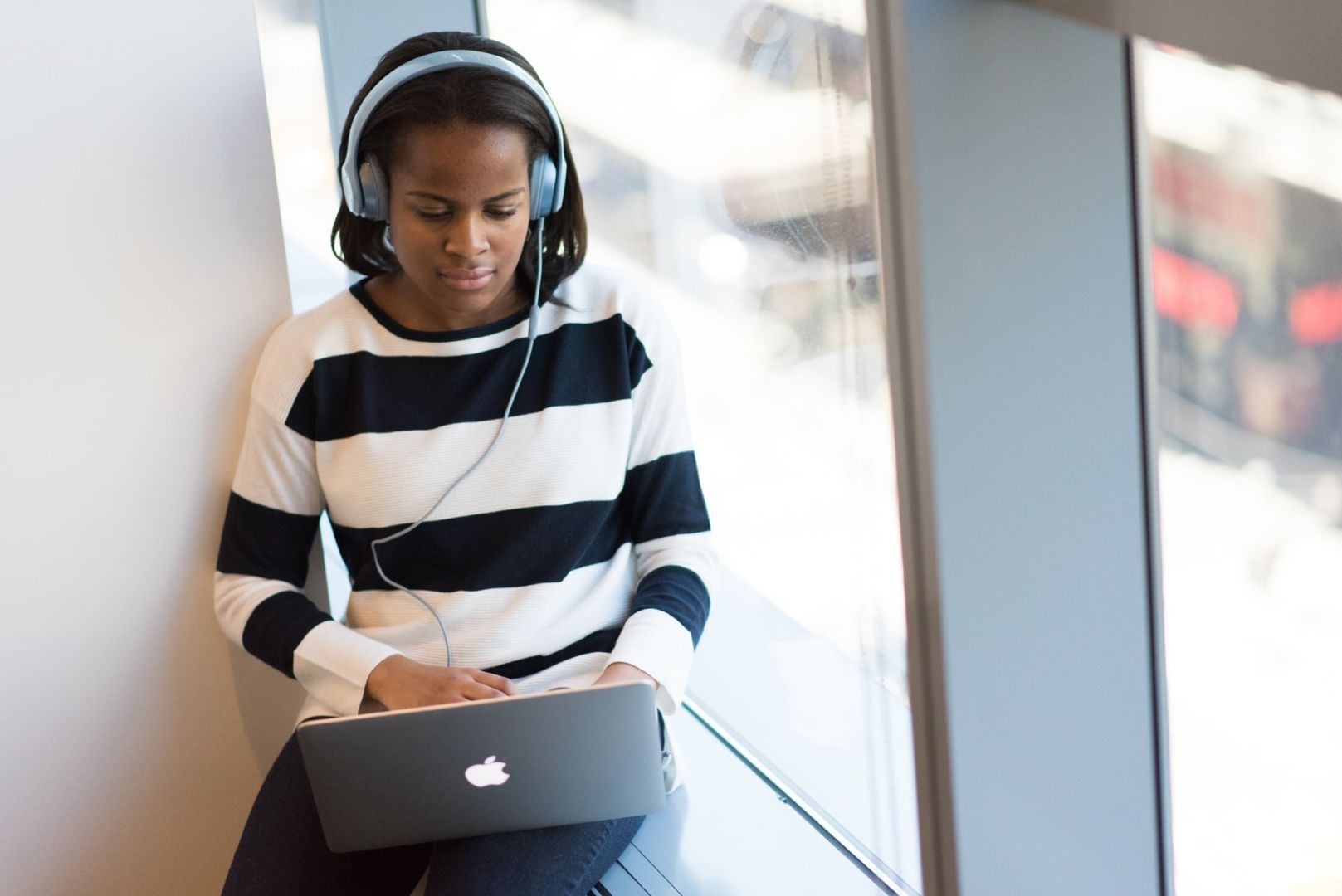 Black woman, sits on window ledge typing on a laptop and she is waring a black and white long sleeve sweater and appears to be listening to music via headphones.