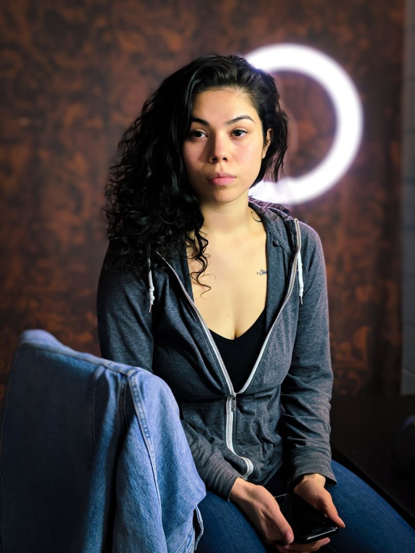 Native American Woman in a hoodie Sitting in front of a ring light and rust colored background, with a jean jacket on the chair back.