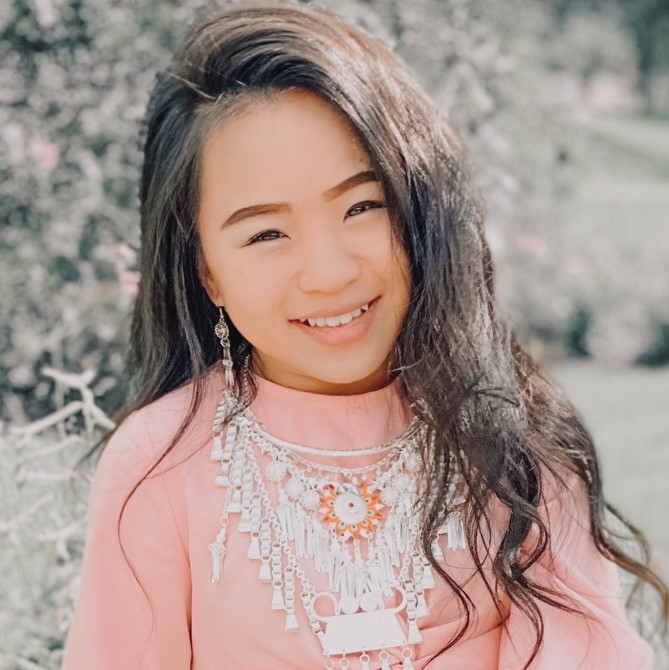 Gaochi sits in the sun, smiling, with light green trees behind her. She has black hair and is wearing a light pink top with a silver traditional Hmong necklace. The necklace has an orange flower pendant in the middle, and Gaochi is also wearing silver Hmong earrings