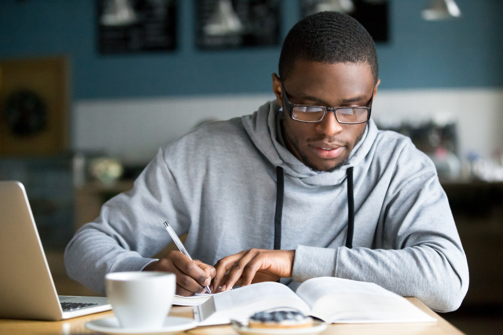 Black man with glasses and wearing a grey athletic sweatshirt. He is reading a book and writing down notes.