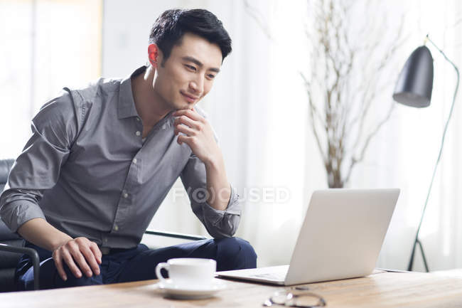 In a minimalist, bright room, with lots of windows that fill the background of the image with bright white light a well-dressed Asian man wearing a gray collared, button down shirt and black pants is sitting on a couch, leaning forward toward his laptop that is on top of his coffee table with his perfect white teacup and saucer. He leans forward with a slight smile, he touches his chin gently with his left hand gently and looks with contemplation into the laptop screen. Off to the right in the background there is a blurred out artsy and simplistic floor plant contrasting darkly against the bright white room.