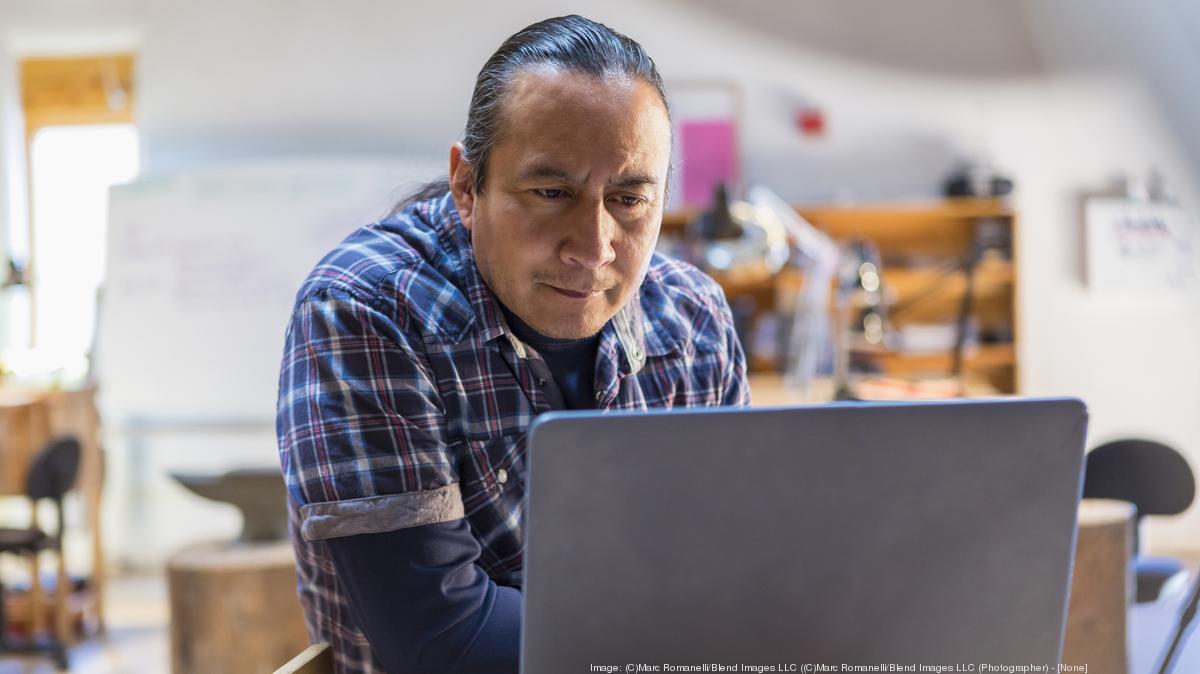 A Native man with long black hair, pulled back in a pony tail, wearing a blue, dark blue and white flannel is sitting at a table,leans slightly forward toward his laptop that is on top of the table, as he stares intensely into his laptop monitor in the library among the blurred stacks in the background.
