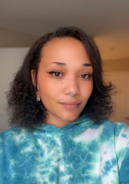[Image Description: Nze is wearing a tie-dye teal/blue hoodie, has short curly hair, has a teal nose ring piercing, is wearing brown dangling earrings and is smiling. There is a white door on the right side behind Nze.] 